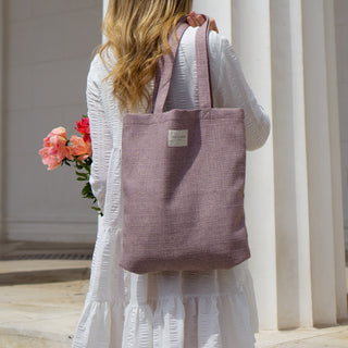 Frau trägt eine Stofftasche Shopper M in der Farbe Lavender Bliss von De La Mur, im Freien vor einer Säule, mit Blumen in der Hand.