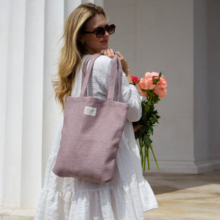 Frau trägt eine Stofftasche Shopper M in der Farbe Lavender Bliss von De La Mur, im Freien vor einer Säule, mit Blumen in der Hand.