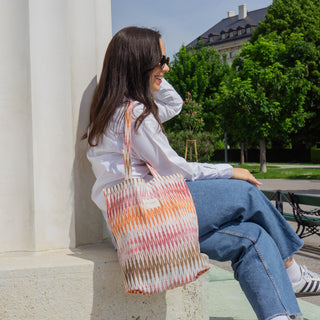 Frau trägt eine Stofftasche Shopper M in der Farbe Sunset Ikat von De La Mur, im Freien vor einer Säule sitzend und lächelnd.