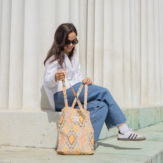 Frau sitzt mit einer Stofftasche Shopper M in der Farbe Vibrant Ikat von De La Mur, im Freien vor einer Säule, lächelnd."