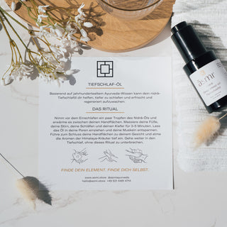 Tiefschlaföl mit dem Duft von Night Jasmine von Asmi Ayurveda, und einem dazugehörigen Infoflyer präsentiert neben dekorativen Blumen auf einem Marmortisch.