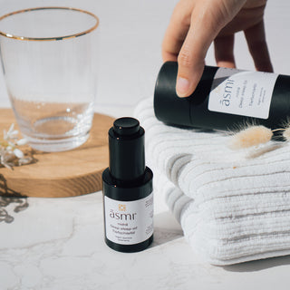 Tiefschlaföl mit dem Duft von Night Jasmine von Asmi Ayurveda, präsentiert neben einem Glas, weißen Handtüchern und dekorativen Blumen auf einem Marmortisch mit einer Hand die nach der Verpackung greift.
