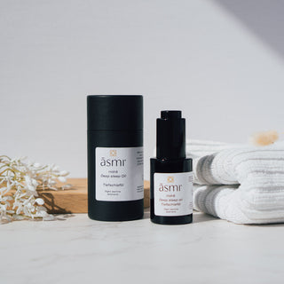 Tiefschlaföl von Asmi Ayurveda mit dem Duft von Night Jasmine, präsentiert in einer schwarzen Flasche und Verpackung neben weißen Handtüchern und dekorativen Elementen auf einem Marmortisch.