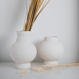Große und kleine Vase Athens von der Marke Claet, handgefertigt mit feinen Rillenstrukturen in elegantem Weiß. Modernes und minimalistisches Design, perfekt als Dekorationsstück oder für Trockenblumen, präsentiert mit zarten Pampasgras-Details.