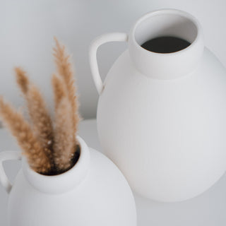 Minimalistische Vase Ghara groß und klein, handgefertigt von Claet. Elegantes Design mit sanft gerundeter Form und praktischem Griff, perfekt für zeitgenössische Dekorationen oder Trockenblumenarrangements. Gefertigt in hochwertigem, strukturiertem Weiß.