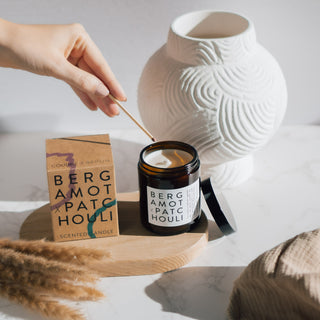Vegane Duftkerze der Sorte Bergamot & Patchouli von Coudre Berlin, in einem braunen Glasgefäß, das mit einem Streichholz angezündet wird, neben der Verpackung auf einem Holztablett mit dekorativen Elementen im Hintergrund.