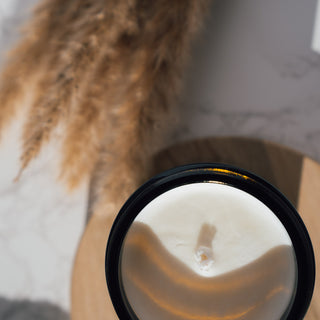 Vegane Duftkerze der Sorte Bergamot & Patchouli von Coudre Berlin, in einem braunen Glasgefäß, von oben dargestellt mit Docht, auf einem Holztablett mit dekorativen Elementen im Hintergrund.