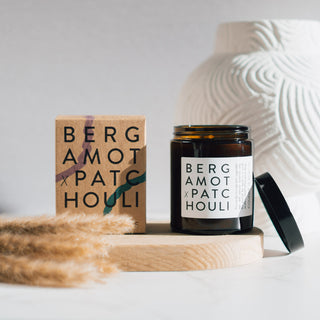 Vegane Duftkerze der Sorte Bergamot & Patchouli von Coudre Berlin, präsentiert in einem braunen Glasgefäß neben der Verpackung auf einem Holztablett mit dekorativen Elementen im Hintergrund.