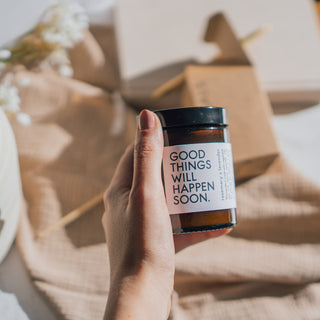 Vegane Duftkerze 'Good Things Will Happen Soon' mit den Duftnoten Rosmarin und Lavendel von Coudre Berlin, wird von einer Person in der Hand gehalten mit weichem Stoff und dekorativen Blumen im Hintergrund.