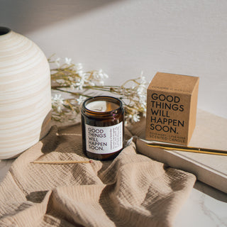 Vegane Duftkerze 'Good Things Will Happen Soon' mit den Duftnoten Rosmarin und Lavendel von Coudre Berlin, präsentiert in einem braunen Glasgefäß neben der Verpackung auf einem weichen Stoff mit dekorativen Blumen im Hintergrund.