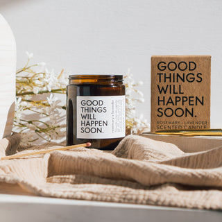 Vegane Duftkerze 'Good Things Will Happen Soon' mit den Duftnoten Rosmarin und Lavendel von Coudre Berlin, präsentiert in einem braunen Glasgefäß neben der Verpackung auf einem weichen Stoff mit dekorativen Blumen im Hintergrund.