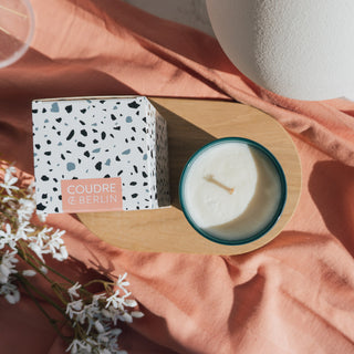 Top-Down-Ansicht der veganen Duftkerze Grapefruit & Cinnamon von Coudre Berlin mit weißem Wachs und Holzdocht, neben der stilvollen Verpackung mit Terrazzo-Muster auf einem Holzpodest, dekoriert mit Stoff und weißen Blüten.