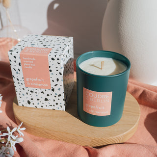 egane Duftkerze Grapefruit & Cinnamon von Coudre Berlin in dunkelgrünem Glas mit sichtbarem Wachs und Holzdocht, neben der passenden Verpackung auf einem Holzpodest arrangiert – umgeben von weichen Stoffen in warmen Tönen.