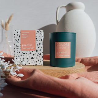 Vegane Duftkerze Grapefruit & Cinnamon von Coudre Berlin im dunkelgrünen Glas mit dazugehöriger Verpackung, dekorativ auf einem Holzpodest arrangiert – umgeben von sanften Stofftexturen und weißen Blüten.