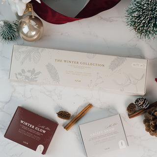 Zwei Seifenstücke aus der limitierten Winterkollektion von Azur Natural Bodycare, präsentiert in stilvollen Verpackungen mit den Namen 'Crispy Snow' (hellgrau) und 'Winter Glow' (dunkelrot). Die Seifen liegen auf einer weißen Marmoroberflächel, weihnachtlichen Dekorationen, Zimtstangen und einem dezenten, winterlichen Ambiente.