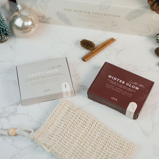 Zwei Seifenstücke aus der limitierten Winterkollektion von Azur Natural Bodycare, präsentiert in stilvollen Verpackungen mit den Namen 'Crispy Snow' (hellgrau) und 'Winter Glow' (dunkelrot). Die Seifen liegen auf einer weißen Marmoroberfläche, begleitet von einem handgefertigten Seifenbeutel aus natürlichem Netzmaterial, weihnachtlichen Dekorationen, Zimtstangen und einem dezenten, winterlichen Ambiente.