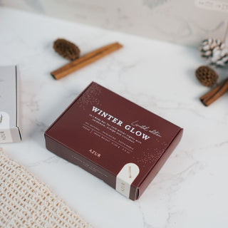 Ein Seifenstück aus der limitierten Winterkollektion von Azur Natural Bodycare, präsentiert in stilvoller Verpackunge mit dem Namen 'Winter Glow' (dunkelrot). Die Seife liegt auf einer weißen Marmoroberfläche, begleitet von einem handgefertigten Seifenbeutel aus natürlichem Netzmaterial, weihnachtlichen Dekorationen, Zimtstangen und einem dezenten, winterlichen Ambiente.