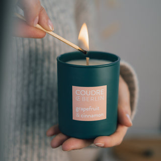 Vegane Duftkerze Grapefruit & Cinnamon von Coudre Berlin wird mit einem Streichholz entzündet. Die Kerze befindet sich in einem dunkelgrünen Glasgefäß und wird in einer Hand gehalten, während die andere das brennende Streichholz an den Docht führt. Eine gemütliche Atmosphäre mit weichem Strickpullover im Hintergrund.