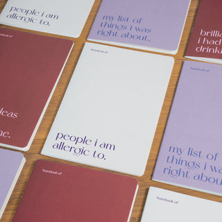 Mehrere Notizhefte im minimalistischen Design, darunter das Notizheft ‘Allergic’ von Typealive mit der Aufschrift ‘people I am allergic to’, arrangiert auf einer Holzoberfläche.