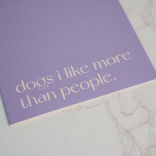 Nahaufnahme des Notizhefts „Dogs I Like More“ in Lavendel mit minimalistischer Typografie auf marmoriertem Hintergrund – ein Must-have für alle Hundeliebhaber.
