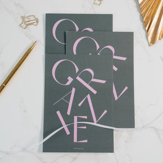 Elegante Postkarte mit der Aufschrift ‘Grazie’ in rosa Schrift auf einem dunklen Hintergrund, arrangiert mit goldenen Büroklammern, einem goldenen Kugelschreiber und dekorativen Elementen, von Typealive.