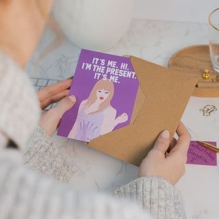 Eine lila Postkarte mit der Aufschrift “It’s Me. Hi. I’m the Present. It’s Me.” wird aus einem braunen Kraftpapier-Umschlag gezogen. Eine handgeschriebene Notiz mit “Geburtstag Sandra” und ein goldener Stift liegen daneben – eine kreative Geburtstagskarte mit musikalischem Bezug.