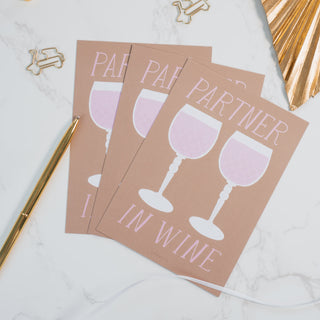 Stilvolle Postkarte mit der Aufschrift ‘Partner in Wine’ und zwei illustrierten rosa Weingläsern auf einem beigefarbenen Hintergrund, arrangiert mit einem goldenen Kugelschreiber und dekorativen Elementen, von Typealive.”