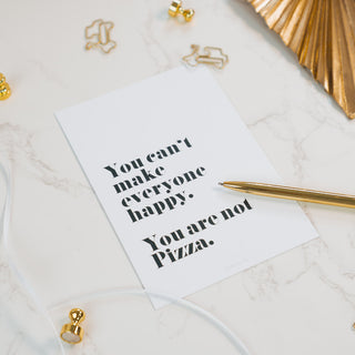 Postkarte mit dem Zitat ‘You can’t make everyone happy. You are not Pizza.’ auf weißem Marmorhintergrund, dekoriert mit goldenen Büroklammern und einem goldenen Kugelschreiber, von Typealive.