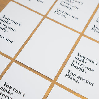 Stilvolle Anordnung mehrerer humorvoller Postkarten mit dem Spruch ‘You can’t make everyone happy. You are not Pizza.’ auf einem Holztisch, in schwarzer Schrift auf weißem Hintergrund, von Typealive.