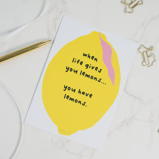 Postkarte mit gelber Zitrone und der humorvollen Aufschrift ‘When life gives you lemons… you have lemons.’ auf weißem Marmorhintergrund, dekoriert mit einem goldenen Kugelschreiber und goldenen Büroklammern, von Typealive.